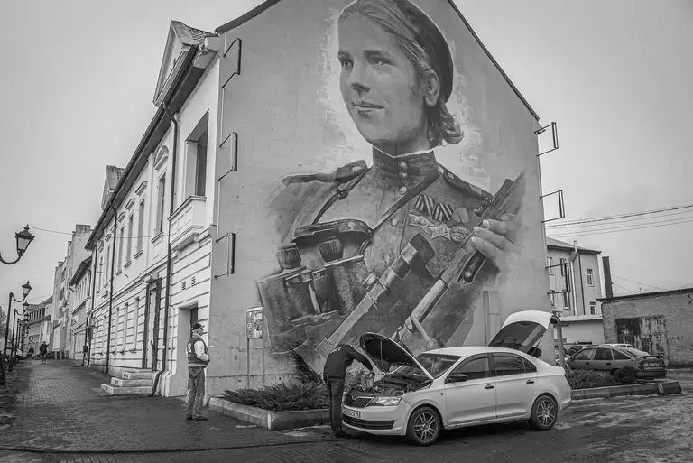 A giant Soviet soldier on the side of a building. Below, a man repairs his car — everyday life under monumental memory. Kaliningrad region, 02.03.2026. Photo by Oleg Klimov